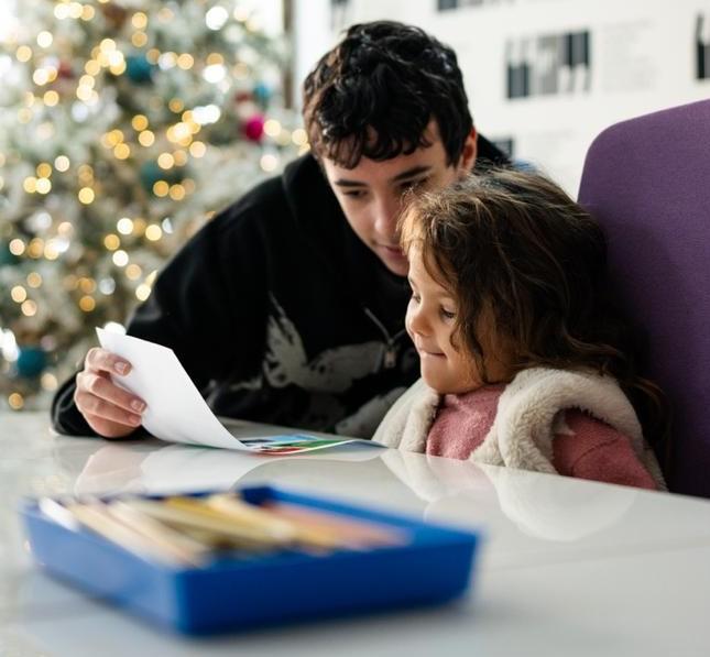Joven y niña leyendo juntas un documento en mesa con material educativo en entorno interior con decoración navideña