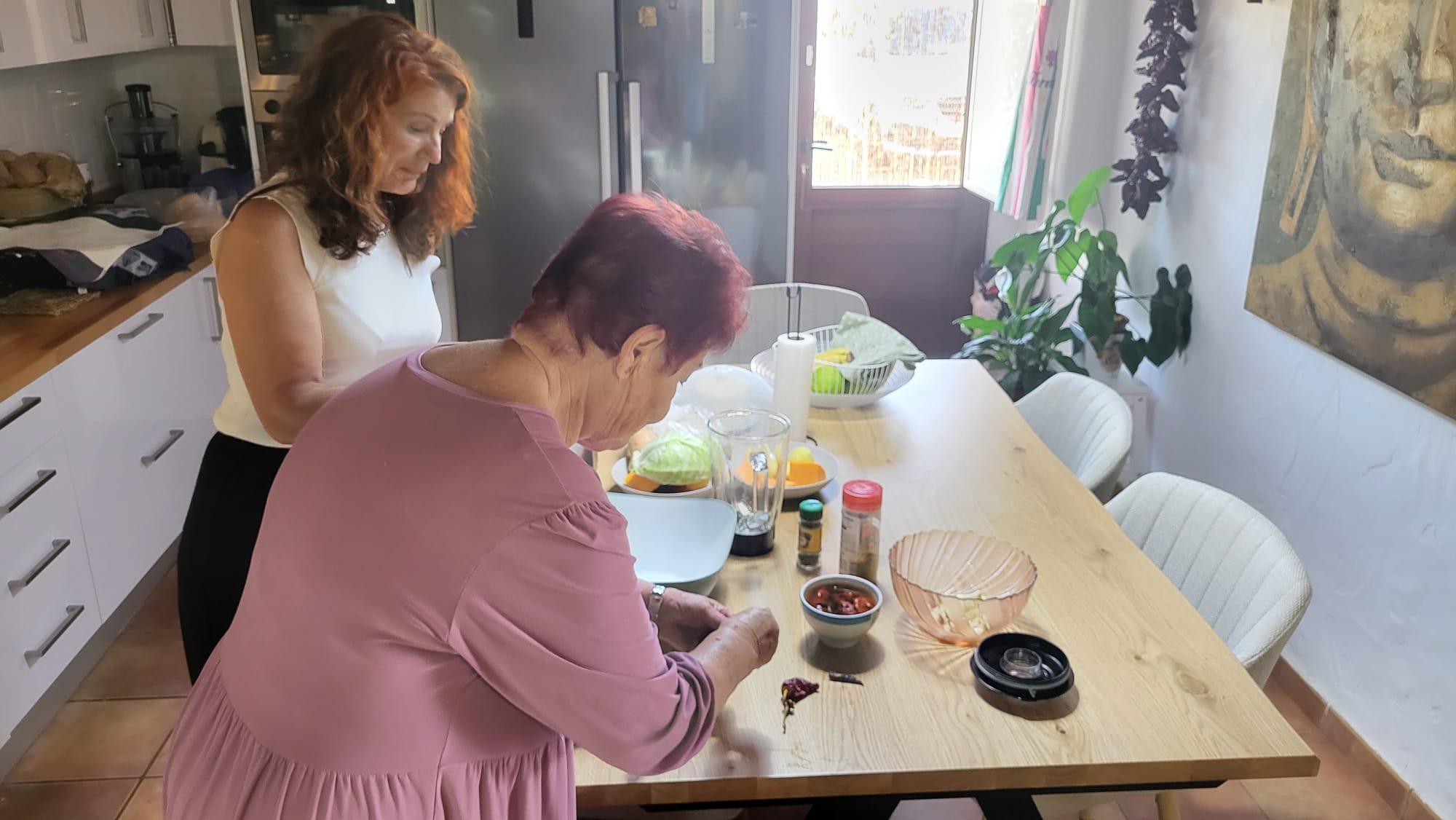 Dos mujeres preparando alimentos en cocina doméstica sobre mesa de madera con ingredientes y utensilios en ambiente cotidiano