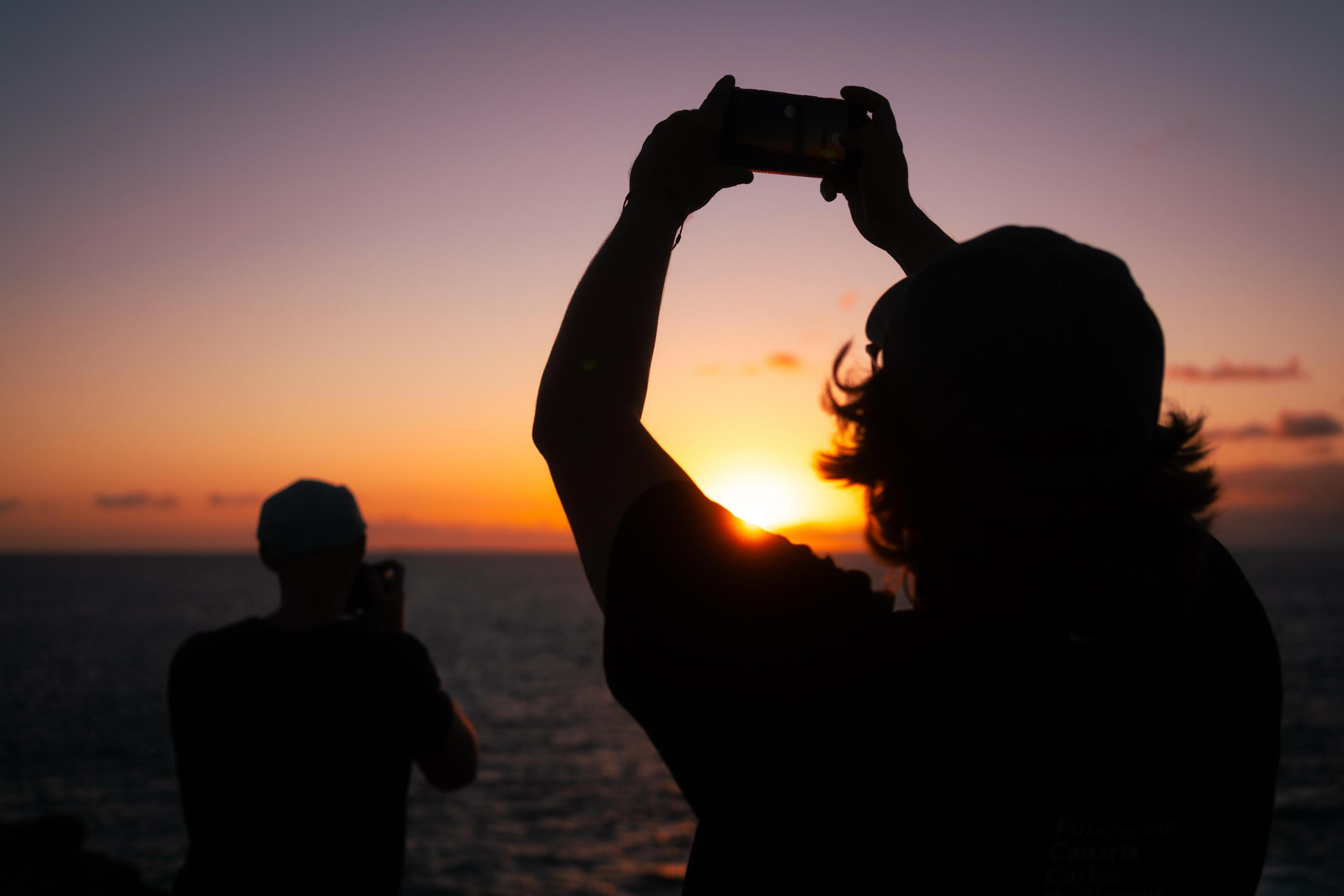 Siluetas de personas fotografiando el atardecer sobre el mar con un móvil frente a un cielo naranja y horizonte