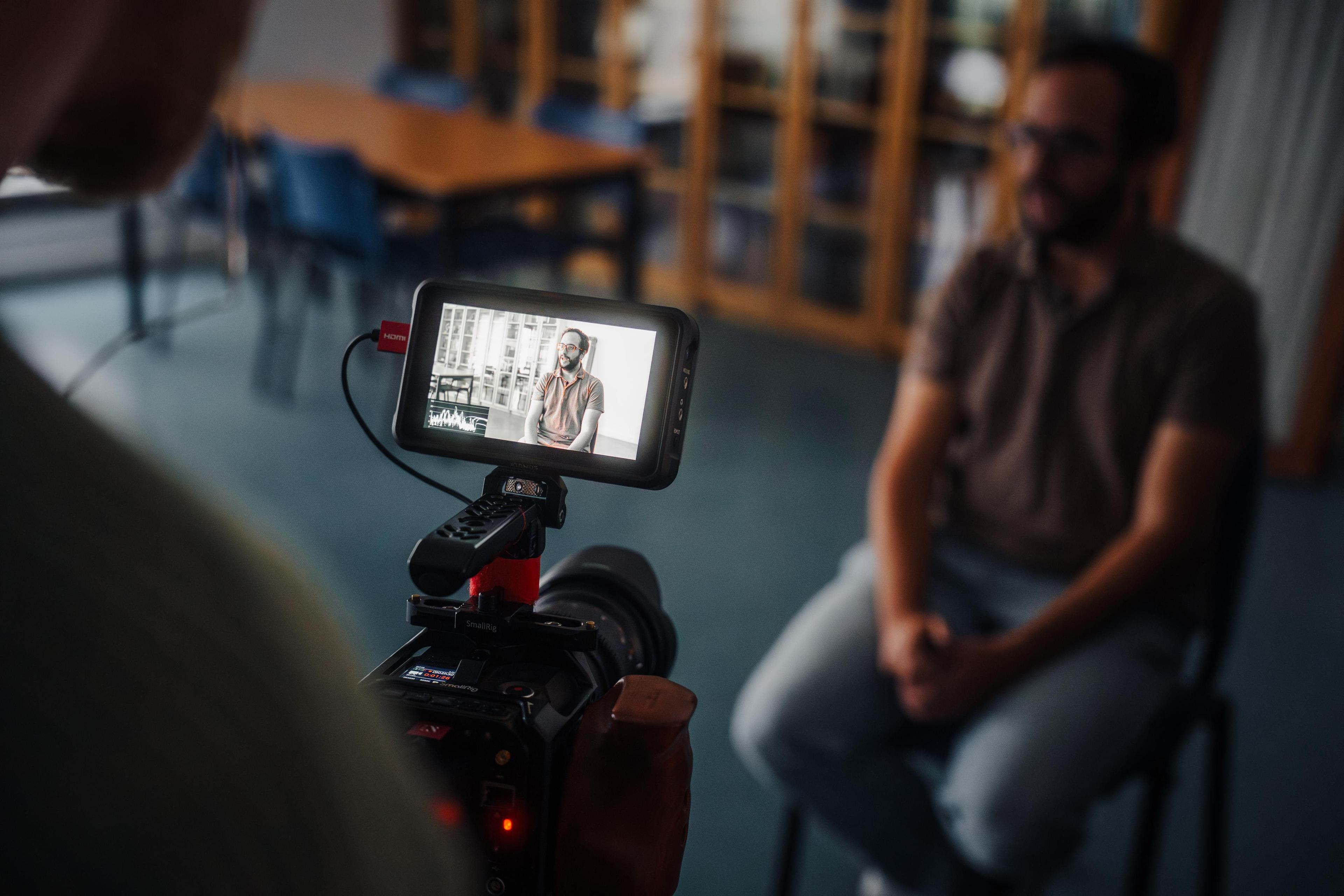 Monitor de cámara grabando entrevista a hombre sentado en interior con estanterías desenfocadas al fondo