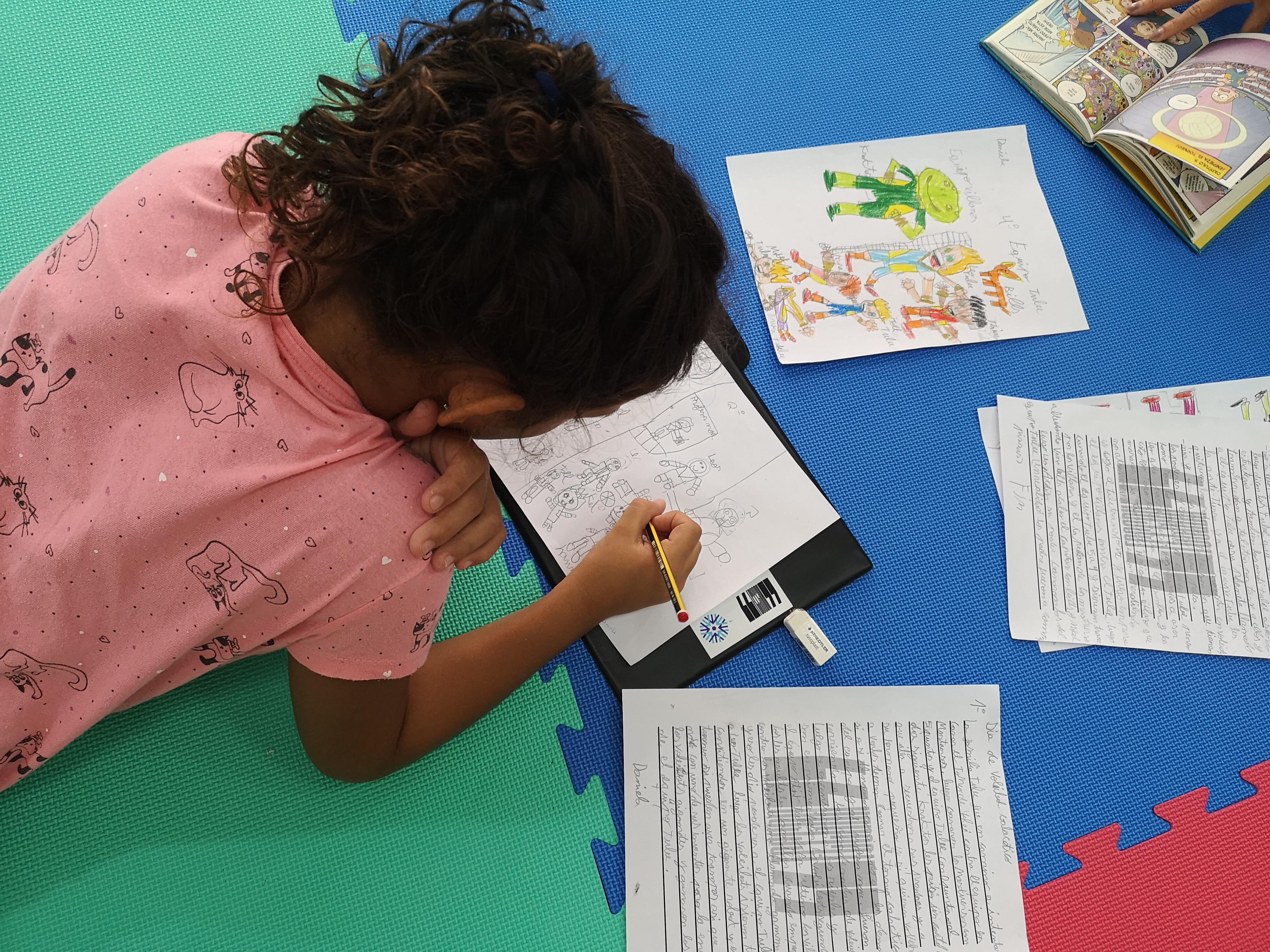 Niña dibujando sobre papel en suelo acolchado con materiales escolares alrededor