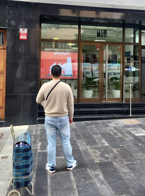 Hombre frente a la entrada de la fundación con carrito de compra, observando el interior a través de puertas acristaladas