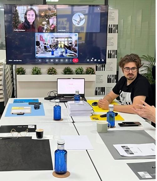 Reunión de trabajo con videollamada en pantalla, participantes sentados en mesa con portátiles y material de oficina