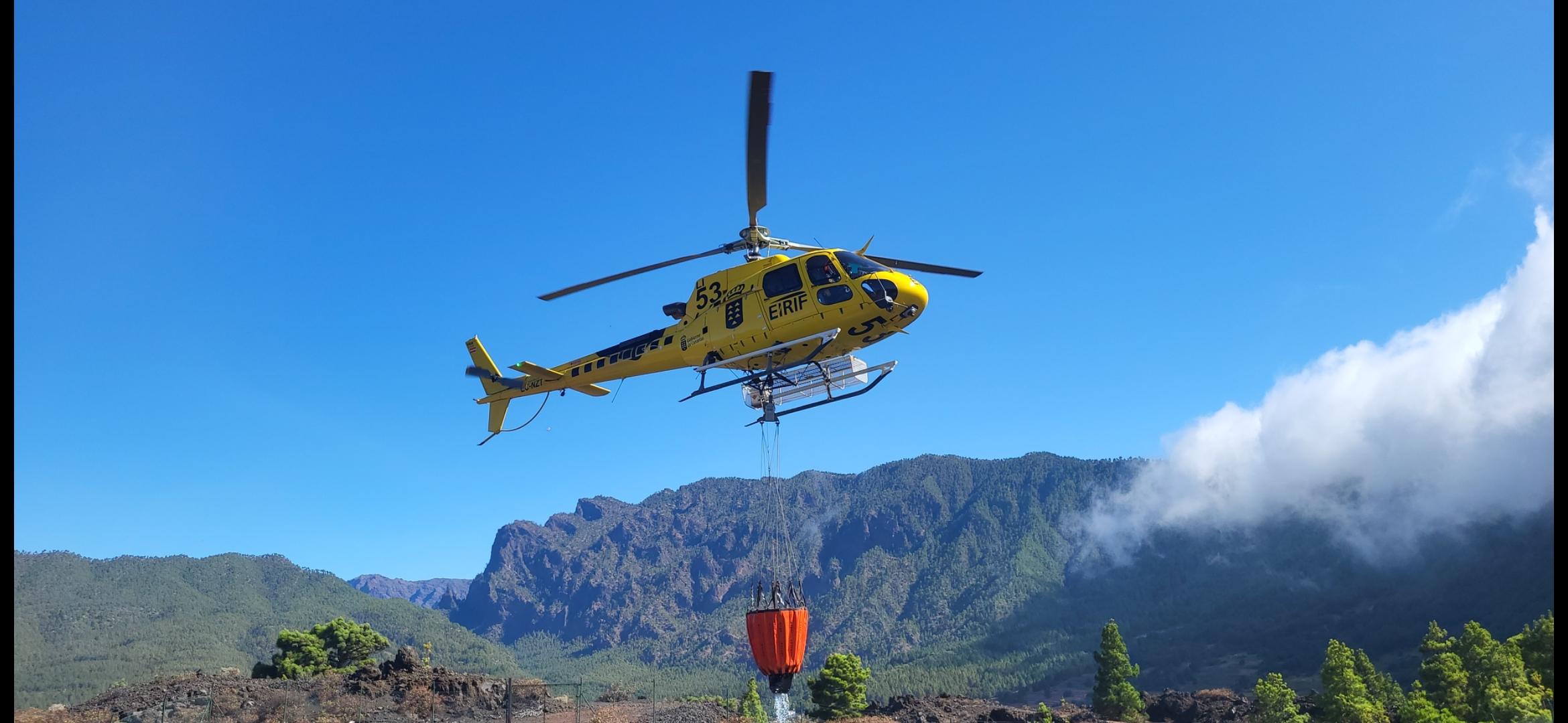 Helicóptero de emergencias sobrevolando paisaje montañoso con depósito de agua colgante en operación aérea