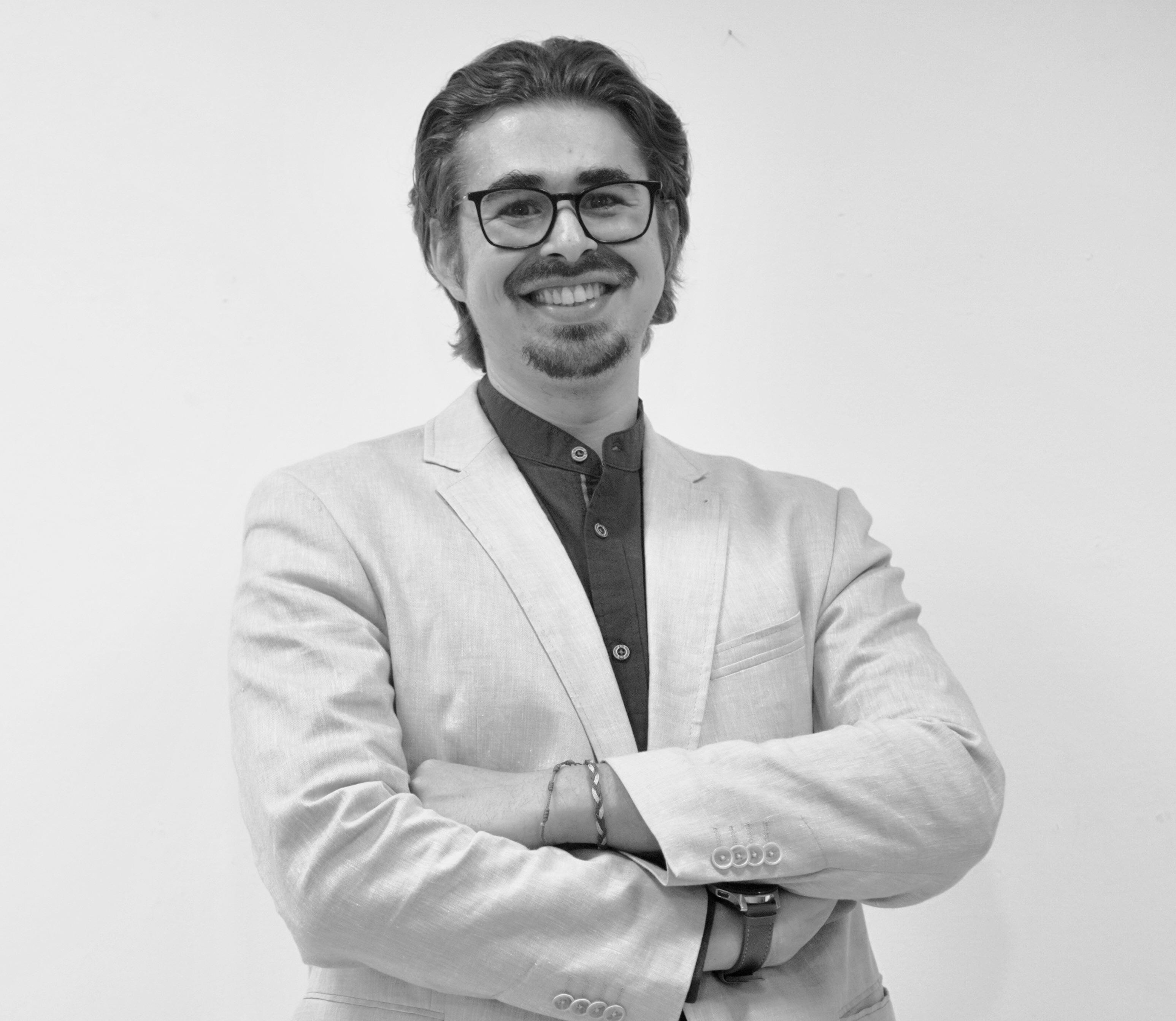 Retrato en blanco y negro de Carlo Brusini Domínguez con gafas y barba, sonriente, brazos cruzados y vestimenta formal clara